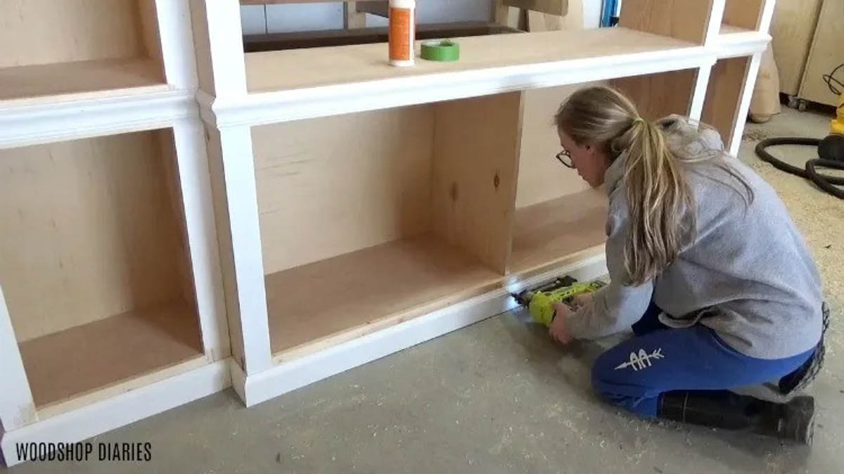 Shara Woodshop Diaries installing baseboard onto middle DIY entertainment center cabinet bottom