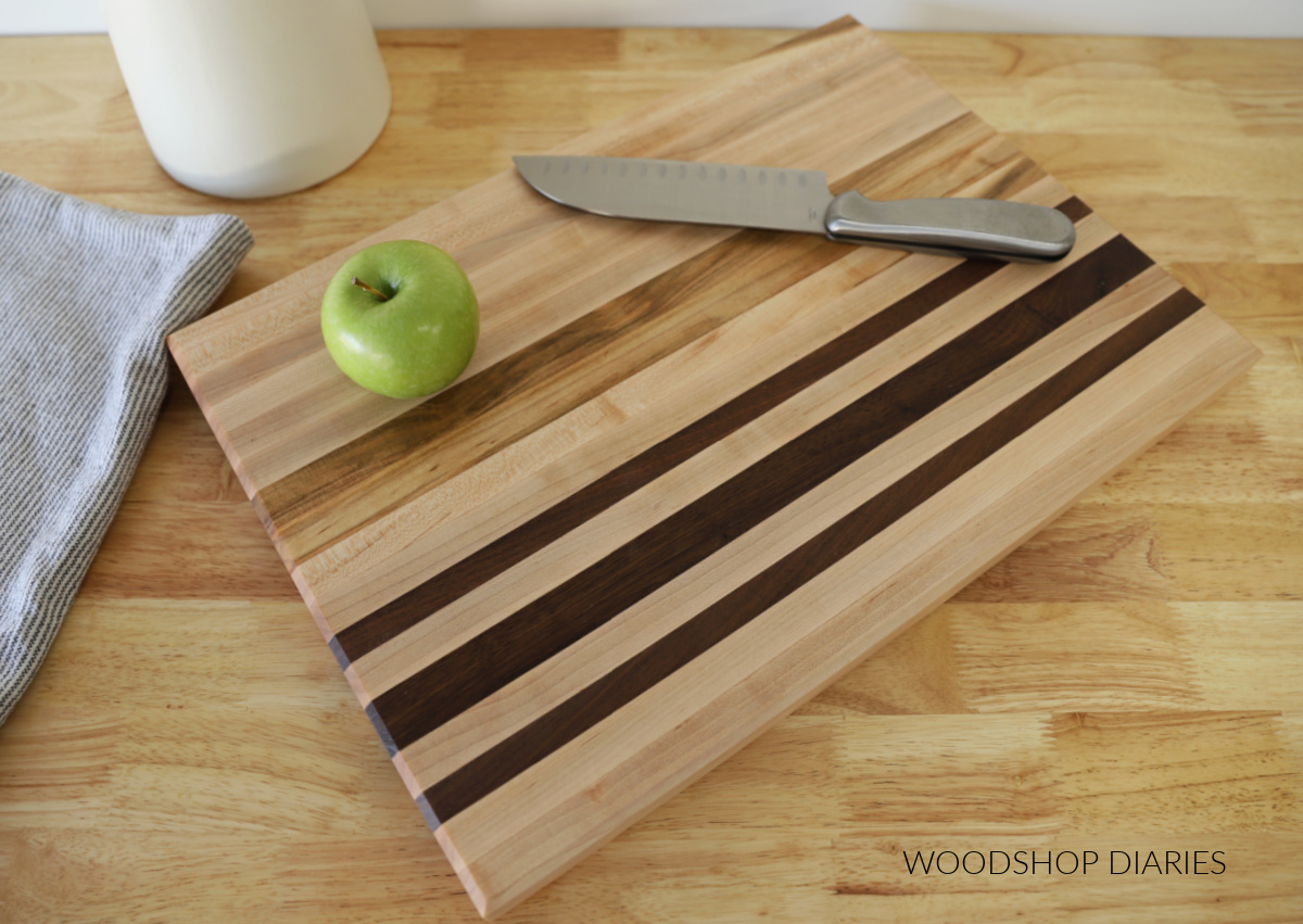 Maple and walnut cutting board laying on butcherblock countertop with green apple and cutting knife on top