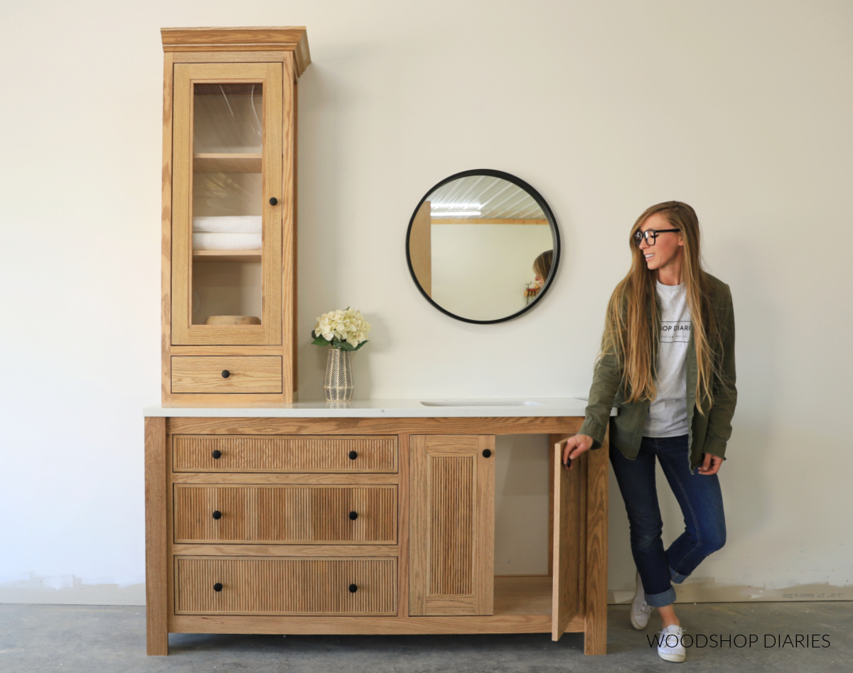 red oak bathroom vanity with off center sink, fluted trim on the drawers and doors and a glass door cabinet on the countertop against white wall