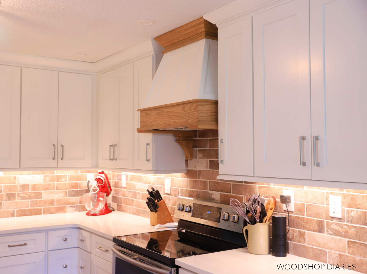 Completed DIY range hood cover installed in kitchen with white cabinets and brick backsplash