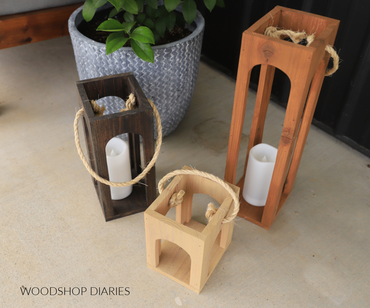 Three wooden lanterns in different sizes stained different colors on porch next to planter
