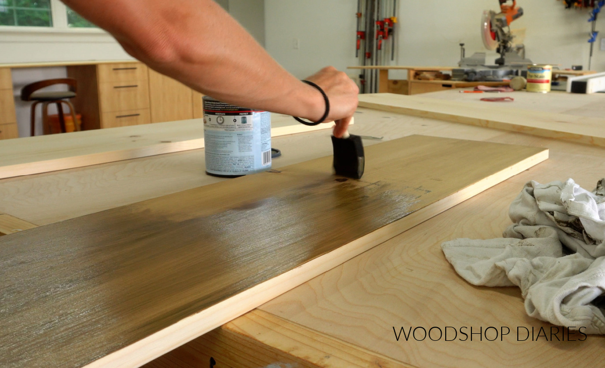 Close up of Shara Woodshop Diaries applying water based stain to 1x12 boards on workbench with foam brush
