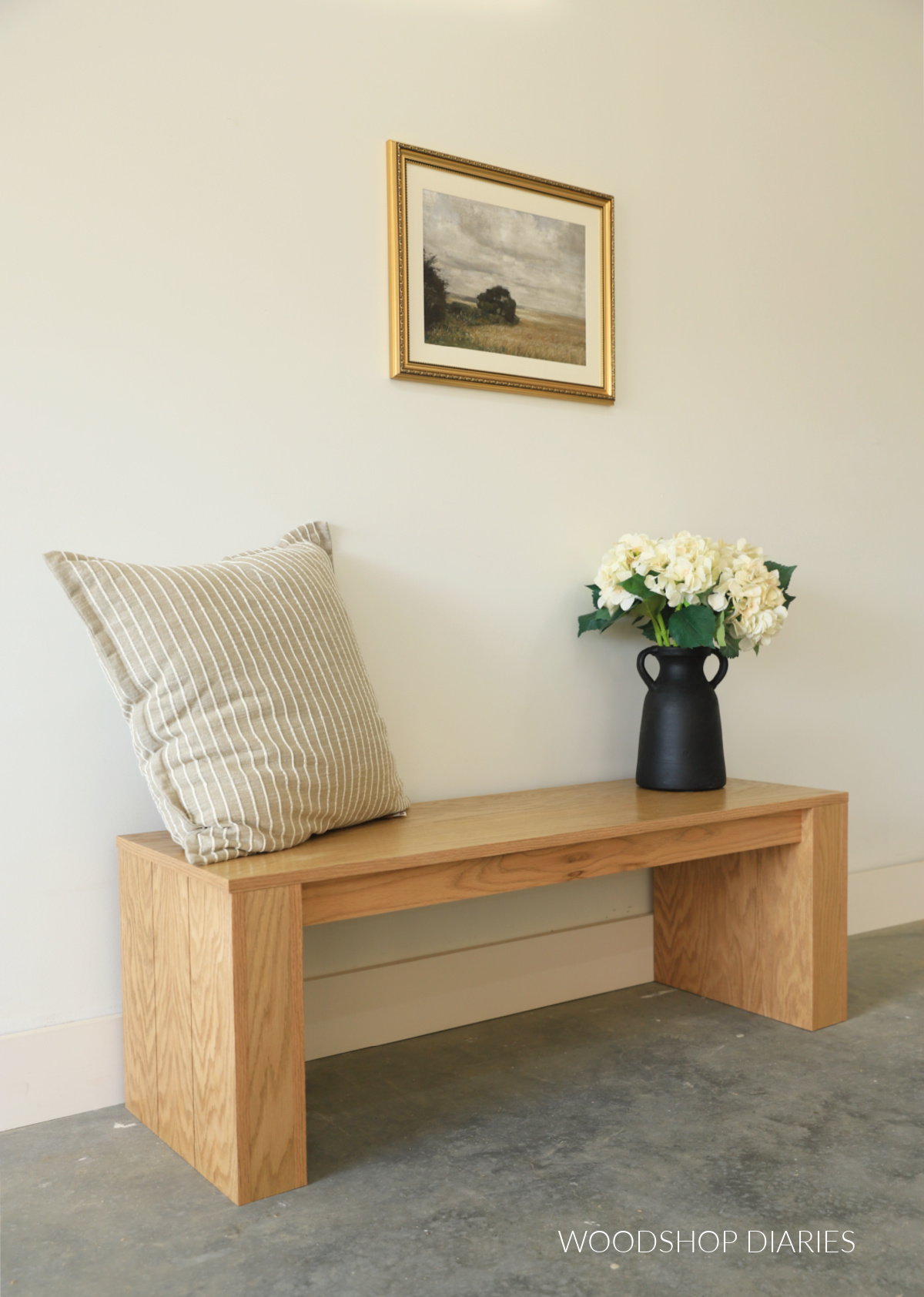 DIY red oak bench made from plywood in front of white wall with pillow, art, and flowers 