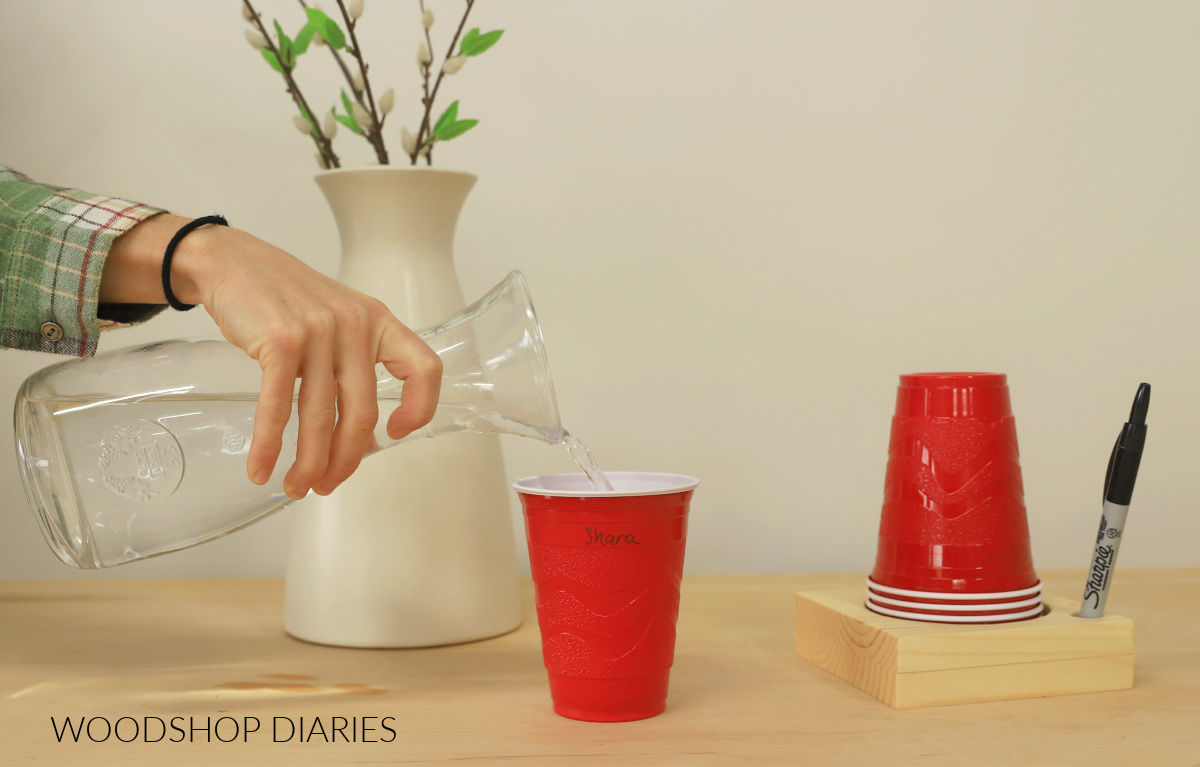 Shara Woodshop Diaries pouring water into red plastic cup. Wood block with red cups stacked and marker resting in hole drilled on side