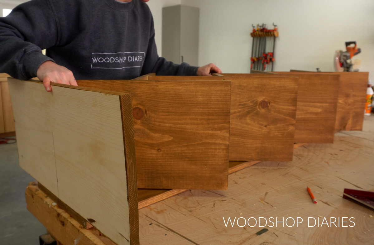 Shara Woodshop Diaries placing shelf assembly on back and bottom boards of bookshelf on workbench